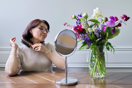 Middle aged woman styling her hair with round comb in front of makeup mirror at home at table. Beauty, hair, care, mature people concept