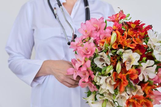 Close-up of bouquet of flowers in hands of female doctor. Medic, pharmacist in white uniform with stethoscope on light background. International, National Doctor's Day, Nurse's Day, Health Day