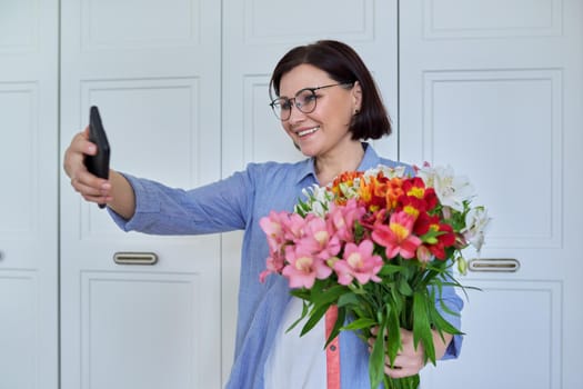 Happy middle-aged woman with bouquet of flowers and smartphone, female taking selfie, talking using video call, on light background at home. Holiday, date, birthday, happiness, joy, mature people
