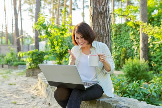 Beautiful emotional middle aged woman relaxing in garden with cup of tea and laptop. Female using video call for virtual meeting, watching movies, reading, having fun