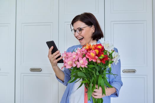 Happy middle-aged woman with bouquet of flowers and smartphone, female taking selfie, talking using video call, on light background at home. Holiday, date, birthday, happiness, joy, mature people