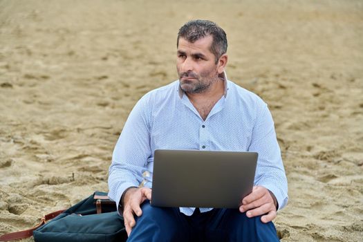 Serious confident mature man with laptop outdoors. Business male working with laptop sitting on sand at beach. Remote business, technology, freelance, lifestyle, leisure, middle age concept