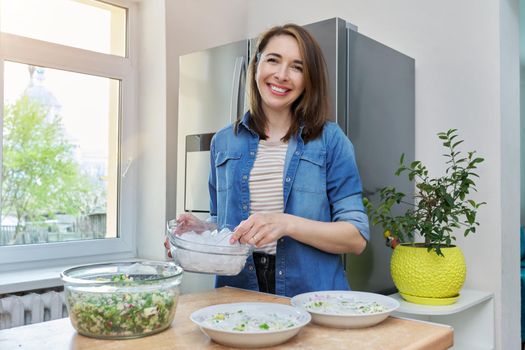 Smiling woman in kitchen near modern chrome large refrigerator with ice for cooling food, vegetable salad in bowl on table. Eating at home, lifestyle, household, dieting, healthy food concept