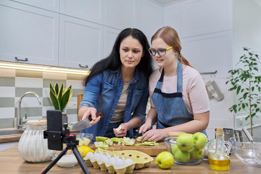 Mom and teen daughter cooking apple pie together, looking at smartphone screen, video recipe, writing recipe on family food blog, talking online using video communication