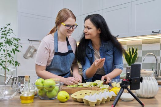 Mom and teen daughter cooking apple pie together, looking at smartphone screen, video recipe, writing recipe on family food blog, talking online using video communication