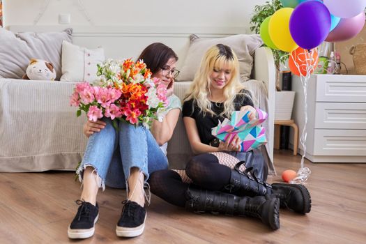 Mother and teen girl, happy birthday. Female mom congratulating daughter with bouquet of flowers and surprise gift box, room interior background. Holiday, celebration, parent teenager relationship