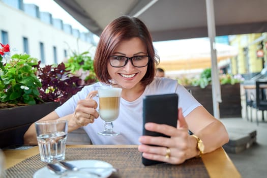 Smiling middle aged woman sitting in an outdoor cafe with cup of coffee and looking at smartphone screen. Coffee break, people 40s age, urban style, leisure, lifestyle, technology concept