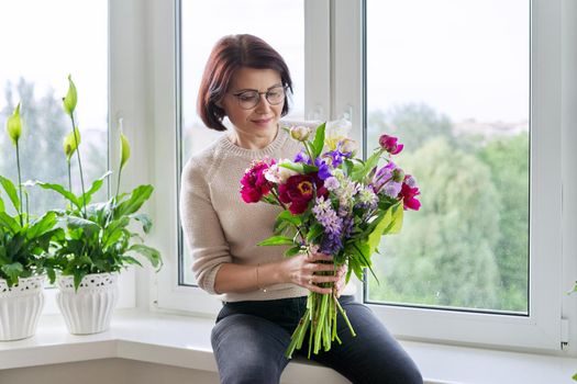 Portrait of beautiful mature woman with bouquet at home. Smiling female with flowers and buds of peony iris lupine. Spring summer season, natural beauty, middle aged people, gift, holiday concept