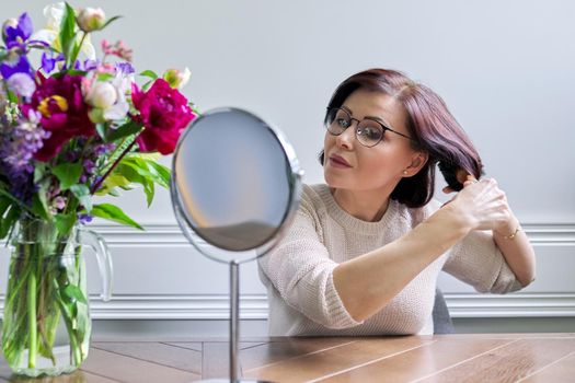 Middle aged woman styling her hair with round comb in front of makeup mirror at home at table. Beauty, hair, care, mature people concept