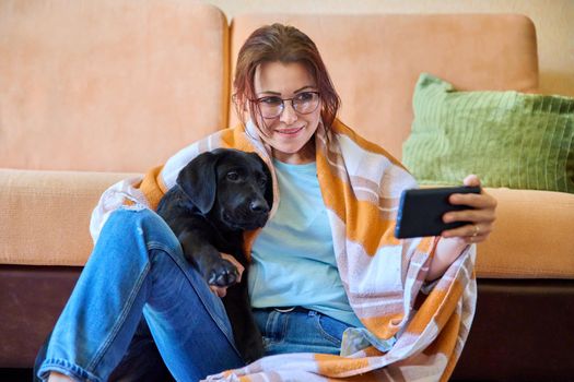 Winter autumn portrait of smiling woman and dog at home. Middle-aged female in warm blanket hugging her pet black labrador puppy takes selfie on smartphone. Lifestyle, love, pets, 40s people concept