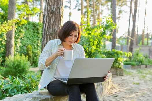 Beautiful emotional middle aged woman relaxing in garden with cup of tea and laptop. Female using video call for virtual meeting, watching movies, reading, having fun