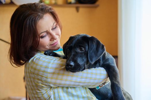 Portrait of middle aged woman and black labrador puppy dog. The owner holding young pet in her arms, in home interior. Lifestyle, love, pets, 40s people concept