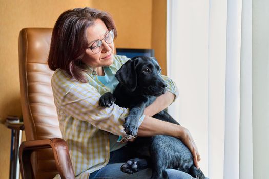 Portrait of middle aged woman and black labrador puppy dog. The owner holding young pet in her arms, in home interior. Lifestyle, love, pets, 40s people concept