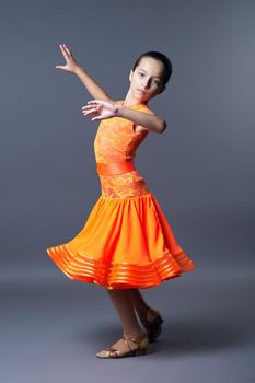 Child girl in an orange sports dress posing in dance movement on gray studio background, sports dances ballroom and latin american for children and adolescents