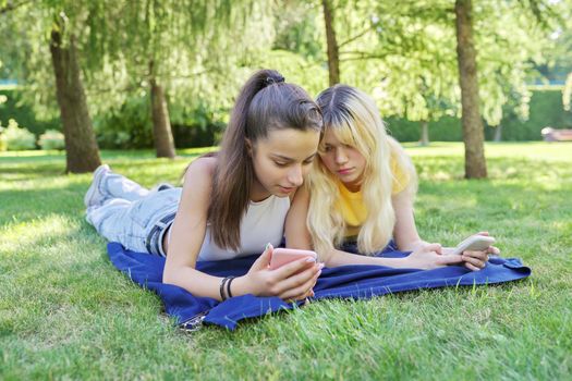 Two young female teenagers lying on grass in park with smartphones. Girls looking at phone screen. Youth, adolescents, technology, lifestyle, leisure concept