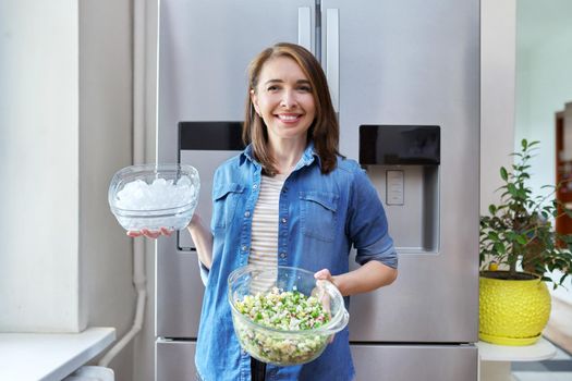 Smiling woman with bowl of ice and plate of vegetable salad in her hands, modern chrome large refrigerator in kitchen. Eating at home, lifestyle, household, dieting, healthly food concept