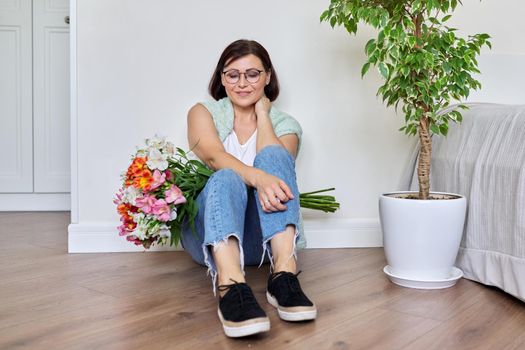 Smiling middle aged woman with bouquet of flowers sitting on the floor at home. Birthday, mother's day, holiday, mature people concept