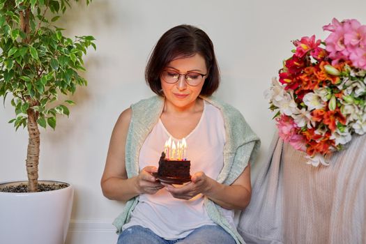 Birthday, celebration, anniversary, 45 years old woman with small cake with burning candles. Smiling middle aged woman with bouquet of flowers sitting on the floor at home.