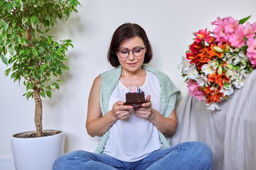 Birthday, celebration, anniversary, 45 years old woman with small cake with candles. Smiling middle aged woman with bouquet of flowers sitting on the floor at home.