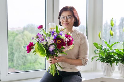 Portrait of beautiful mature woman with bouquet at home. Smiling female with flowers and buds of peony iris lupine. Spring summer season, natural beauty, middle aged people, gift, holiday concept