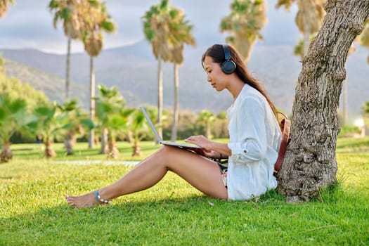 Young woman in headphones working on laptop outdoors. Asian female university student studying remotely, creative freelancer, business woman, blogger vlogger, work and study online, modern technology