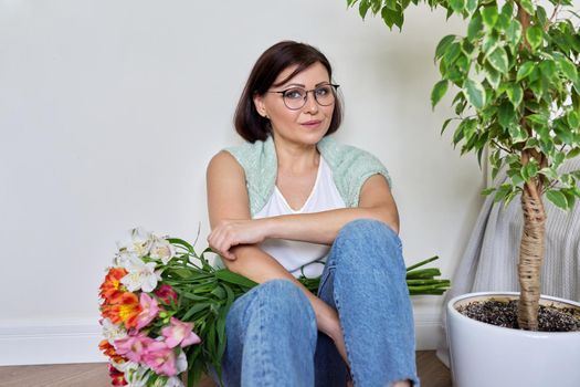 Smiling middle aged woman with bouquet of flowers sitting on the floor at home. Birthday, mother's day, holiday, mature people concept