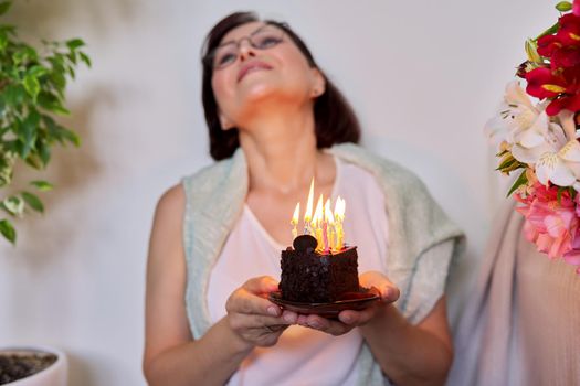 Middle-aged women with small birthday cake with candles. Happy female at home on the floor with bouquet of flowers. Age, date, aging, celebration, lifestyle, anniversary concept