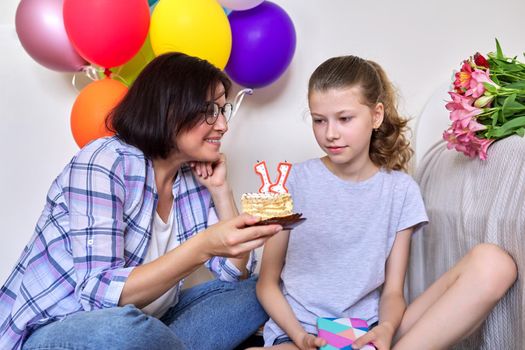 Childs birthday, 11 years old. Mom and daughter sitting together at home with small cake with candles, gift, balloons, bouquet of flowers