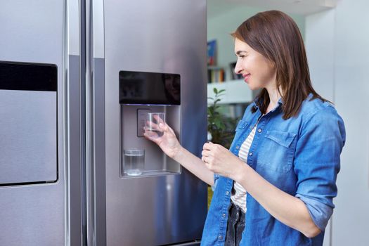 Water dispenser, woman taking cold water into glass from home modern chrome refrigerator in kitchen, dispenser of fridge. Cool water, household, comfort concept