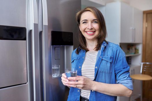 Water dispenser, woman taking cold water into glass from home modern chrome refrigerator in kitchen, dispenser of fridge. Cool water, household, comfort concept