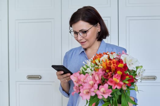 Happy middle-aged beautiful woman with large bouquet of flowers and smartphone, on light background at home. Holiday, date, birthday, happiness, joy, mature people concept