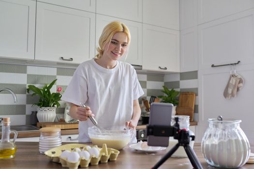 Female teenager learns to cook pancakes, watching video recipe on smartphone, in home kitchen. Cooking home food, culinary hobby, learning cooking class, teenagers, people concept