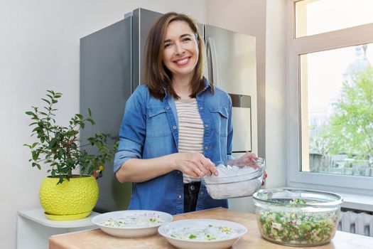 Smiling woman in kitchen near modern chrome large refrigerator with ice for cooling food, vegetable salad in bowl on table. Eating at home, lifestyle, household, dieting, healthy food concept