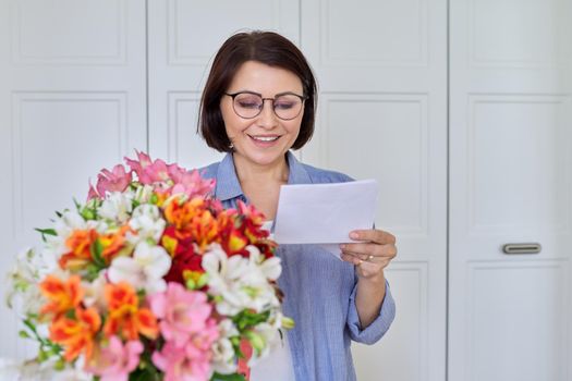 Happy middle aged woman with bouquet of flowers reading text from paper card. Congratulations, date, surprise, date, anniversary