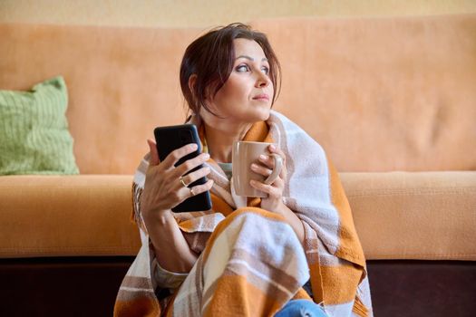 Autumn winter portrait of middle aged woman under warm woolen blanket with hot cup of tea and smartphone in hands. Female resting at home, sitting on the floor near the sofa