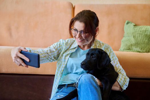 Middle-aged smiling happy female labrador puppy owner taking selfie photo on smartphone with dog sitting together at home on floor