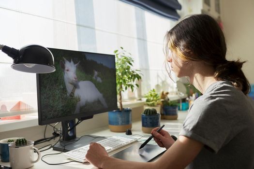 Teen guy sitting at table at home working on computer, using graphics tablet to work with images. Modern technologies, hobby of male teenager, freelance, youth concept