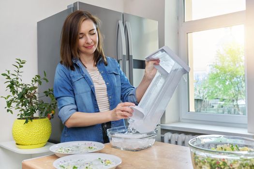 Woman taking ice cubes into bowl in kitchen, modern refrigeration equipment, home chrome refrigerator background