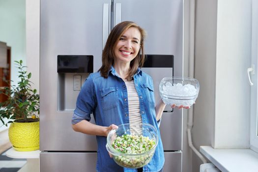 Smiling woman with bowl of ice and plate of vegetable salad in her hands, modern chrome large refrigerator in kitchen. Eating at home, lifestyle, household, dieting, healthly food concept