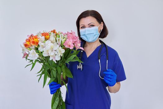 Doctor's Day, Nurse's Day. Female medic in blue uniform medical protective face mask gloves with stethoscope with large bouquet of flowers on light background