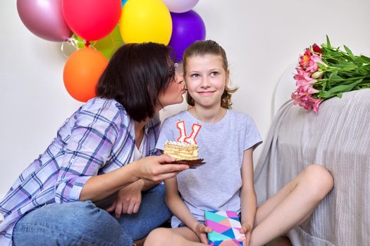 Childs birthday, 11 years old. Mom and daughter sitting together at home with small cake with candles, gift, balloons, bouquet of flowers