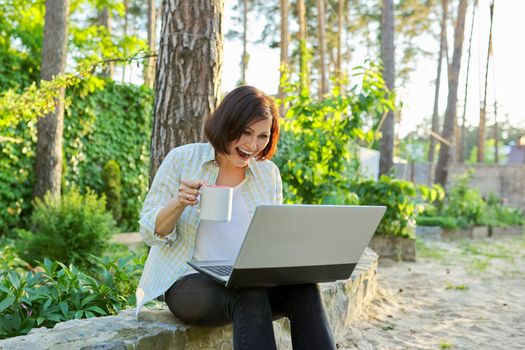 Beautiful emotional middle aged woman relaxing in garden with cup of tea and laptop. Female using video call for virtual meeting, watching movies, reading, having fun