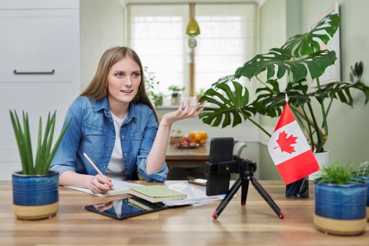 Online learning, distance lessons, e-learning, teenage female university student sitting at home looking at smartphone screen, video meeting, on table canadian flag. Canada, education, study concept