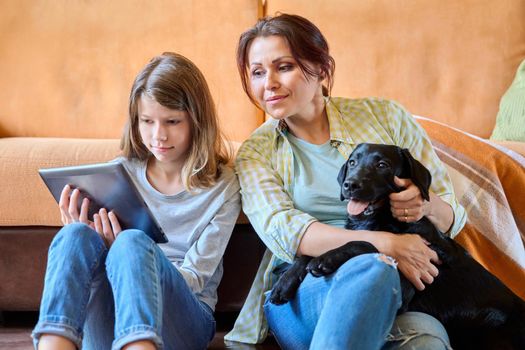 Family, lifestyle, communication, happiness, vacation together, pets, home concept. Mother daughter child and labrador puppy dog sitting together near sofa looking into digital tablet screen at home