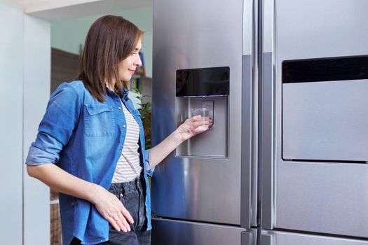 Water dispenser, woman taking cold water into glass from home modern chrome refrigerator in kitchen, dispenser of fridge. Cool water, household, comfort concept