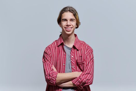 Portrait of smiling guy teenager with crossed arms, hipster teen male 15, 16 years old with long hair looking at camera on light background
