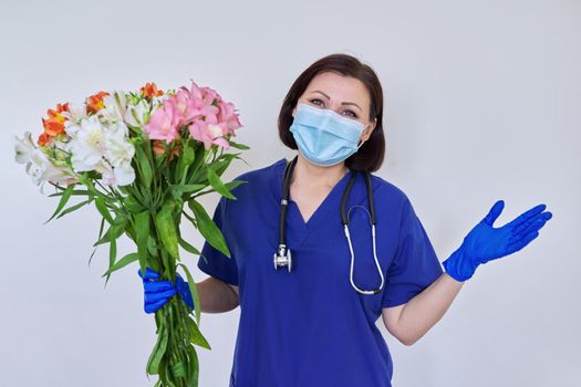 Doctor's Day, Nurse's Day. Female medic in blue uniform medical protective face mask gloves with stethoscope with large bouquet of flowers on light background