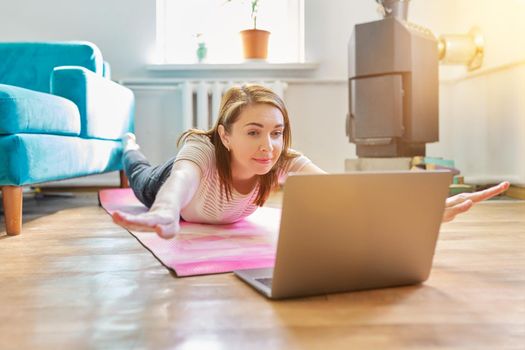 Middle-aged woman doing exercises at home on floor. Female in casual clothes on exercise mat with laptop, online training, healthy active lifestyle