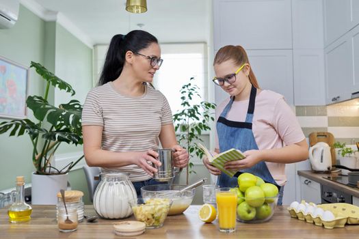 Mom and teenage daughter preparing apple pie in kitchen together, looking at recipe book, talking and laughing. Communication between teenager and parent, lifestyle, cooking at home concept