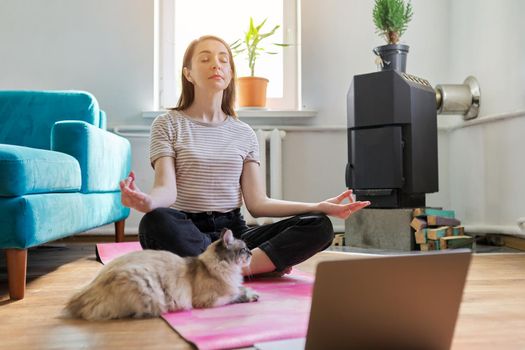 Lifestyle leisure meditation. Middle aged woman sitting at home on floor with laptop in lotus position, pet cat on exercise mat with owner